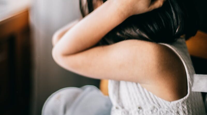 Side view of stressed unrecognizable little girl with black hair sitting at round table and touching head