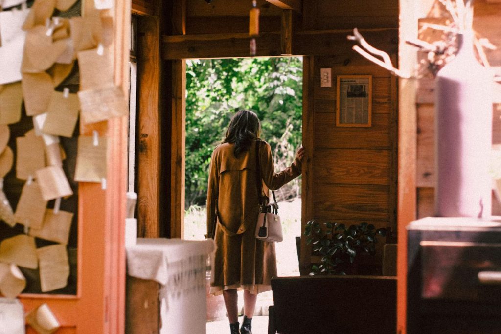 A woman in a warm coat steps out of a cozy cabin into the sunlight. De la violencia machista se sale. 