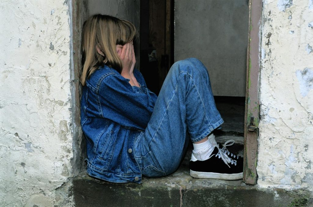 A young girl in denim sits alone in a doorway looking distressed, conveying loneliness. Niño sufre violencia machista