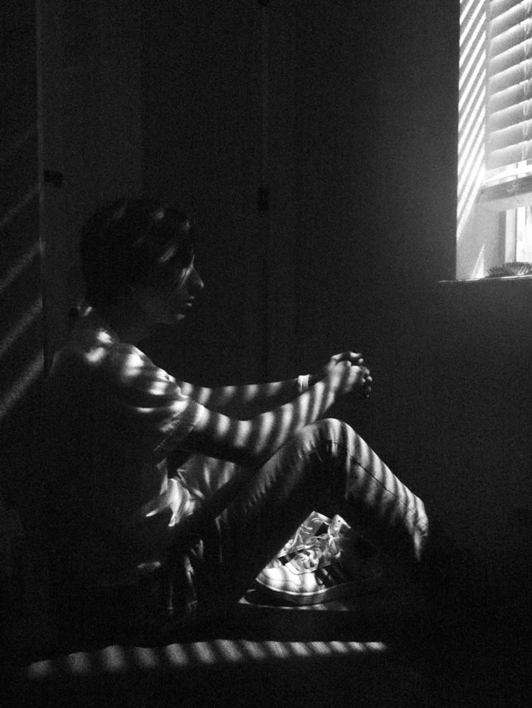 Black and white photo of a thoughtful man sitting by a window with blinds casting shadows. Consecuencias de la violencia machista