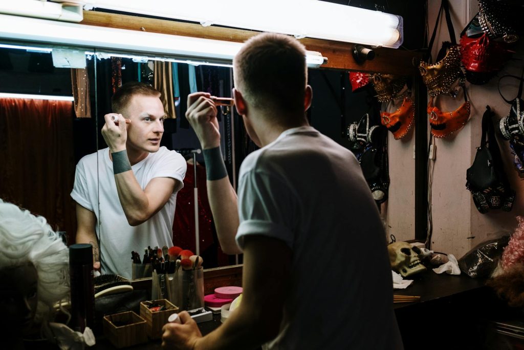 Drag performer applies makeup in a theater dressing room, reflecting transformation and identity. Género